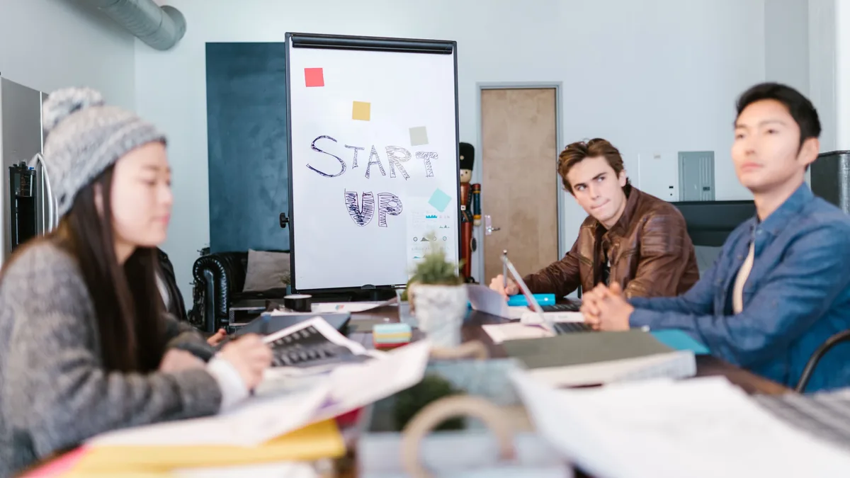 Three people are seated at a table in an office setting, with a whiteboard displaying the words "START UP" in the background.